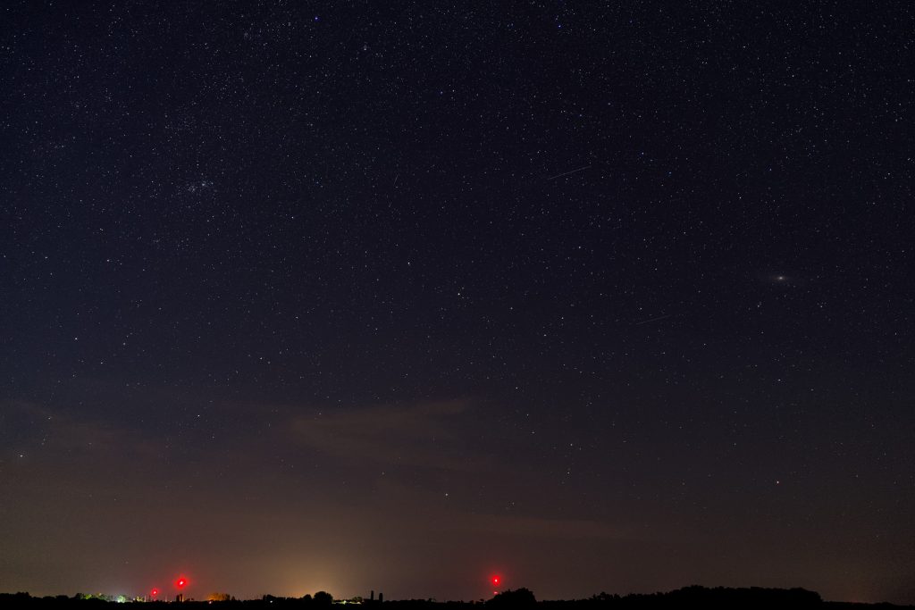 The red lights of radio towers glow on the horizon below a thin layer of clouds. The Double Cluster is visible in the clear sky high above the two radio towers on the left. Meanwhile, the Andromeda Galaxy appears as a bright smudge near the center right.
